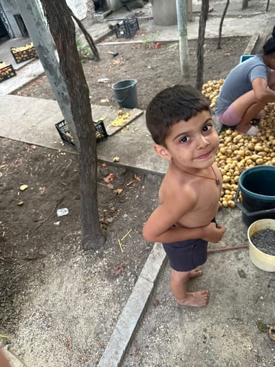 The image depicts a young boy, appearing to be around five to seven years old, standing barefoot on a dirt and concrete surface in what appears to be a rural or outdoor setting, potentially in Mlekarevo, Bulgaria. The boy is shirtless, wearing dark shorts, and looking directly at the camera with a slight smile. He has dark hair and brown eyes and is wearing a red cord necklace with a small pendant.
Behind the boy, to the right, a large pile of potatoes is visible, and a woman in a grey t-shirt and pink shorts is crouching, appearing to sort or handle the potatoes. Two buckets are also present; one dark blue or black, and a lighter colored one filled with small grey stones or gravel.
In the background, there are more crates of what appear to be potatoes, as well as some scattered debris and foliage. A tree trunk is prominent in the left and center of the frame. The overall environment suggests an agricultural or outdoor work area, possibly a backyard or a farm-related space. The lighting indicates it is daytime, with shadows present.