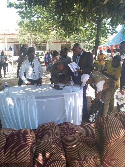 The image depicts a group of men gathered around a white table draped in a tablecloth. The men are in the process of signing a document. There is a black briefcase and two bottles of water on the table. The men are standing in front of a building. There are multiple vehicles parked in the background. The image was taken in Bunia, Democratic Republic of the Congo, at the coordinates 1.557 latitude and 30.244 longitude.