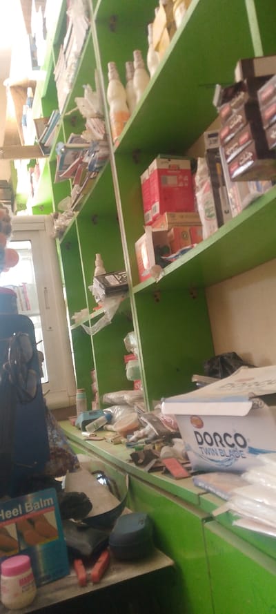 The image displays an interior view of a retail establishment in Danbare, Nigeria. A multi-tiered shelving unit, rendered in a distinct vibrant green, occupies the majority of the frame, extending from the foreground to the background on the right. The shelves are populated with a diverse array of merchandise, including numerous white-colored bottles with patterned caps, various small packaged goods, items encased in clear plastic bags, and stacked cardboard boxes, some red and others brown/yellow. A display rack holding smaller packaged items is visible on the upper right shelf. Below the shelving, a green counter surface is cluttered with assorted items. Identifiable objects on the counter include a white container labeled "DORCO TWIN BLADE," a blue box marked "Heel Balm," a small pink-lidded jar, red-handled pliers, and a dark blue lidded container, intermingled with various smaller, less distinct objects. A partially visible white-framed opening, consistent with a door or window, is present on the far left. No individuals are depicted, and no active interactions or actions are occurring within the visible scene.