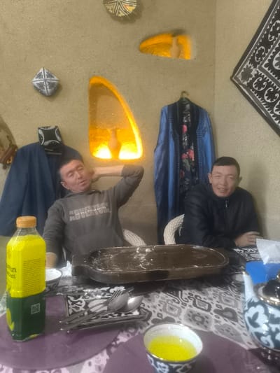 The image captures an indoor scene in Turkkishlak, Uzbekistan, featuring two adult males seated at a table. The room's walls are a light tan, textured, and adorned with several decorative elements: two irregularly shaped, glowing recessed niches emitting warm orange light, two round, patterned wall plates, and a square fabric hanging with geometric patterns. Two traditional robes are displayed on hangers on the wall, one blue and one black with floral designs; a black and white skullcap rests on the blue robe.
At the center of the table, which is covered by a black and white patterned tablecloth, sits a dark brown, oval-shaped wooden serving tray containing white residue, possibly flour or dough. To the left foreground, a tall plastic bottle with a yellow cap and green base holds a yellow-green liquid, alongside a small white bowl and a collection of cutlery on a rectangular dish. To the right foreground, a white teapot with blue and white intricate patterns and gold accents is visible, next to a matching patterned cup containing a yellow liquid. A blue tissue box with white tissues is also present on the table.
The male on the left, wearing a grey hooded sweatshirt with "THE WILD SIDE OF NATURE" text, is seated in a white wicker-backed chair, leaning back with his right hand placed behind his head. His eyes are partially closed, and his mouth is slightly open. The male on the right, dressed in a dark jacket, sits upright, his hands clasped on the table, and smiles directly towards the camera. Both individuals appear to be of East Asian or Central Asian descent.