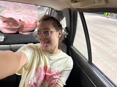 In the city of Jer-Kazar, Kyrgyzstan, a young woman with brown hair pulled back and wearing clear-rimmed glasses is taking a selfie from the back seat of a car. She is sticking her tongue out playfully. She is wearing a light yellow t-shirt with a pink graphic print. In the background, a pink duffel bag is visible on the seat behind her. Through the car window, a paved area with orange cones and a glimpse of a structure with green and yellow accents can be seen, suggesting an outdoor public space.