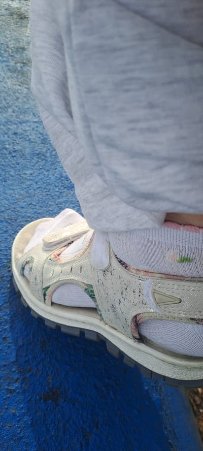 A close-up shot focuses on a person's foot wearing a white sandal with a floral pattern and white socks. The sandal is resting on a textured blue surface. A light grey garment is draped over the foot and sandal, partially obscuring the view. The scene appears to be outdoors, possibly in a park or playground, given the textured blue surface, which could be playground surfacing. There are no specific landmarks or architectural features visible to determine the exact location within Castlebar, Ireland. The lighting suggests daylight.