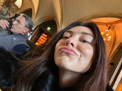 The image depicts an indoor scene in Cernay-la-Ville, France, featuring two individuals within an ornately decorated setting. In the foreground, a female with long dark hair, wearing a black textured garment, is positioned close to the camera, directing a pouty expression with pursed lips and slightly puffed cheeks towards the lens. To her left, an older male with short grey hair and clear-framed eyeglasses is visible from the chest up. He wears a grey sleeveless vest over a dark long-sleeved shirt. His head is turned to his left, and he gazes upward, away from the camera, with his right hand raised near his face.
The background displays classical architectural elements, including arched doorways and vaulted ceilings with decorative moldings. A chandelier with multiple light sources emits warm illumination from the upper right. Through a background archway, patterned wallpaper is visible, along with an illuminated shelving unit containing bottles, and a green exit sign above a doorway. Christmas-themed decorations, featuring red berries and green foliage, are present on the far left. A third individual is partially visible standing in the deeper background near the shelves. The subjects appear to be seated on dark upholstered furniture within a dimly lit, warm-toned interior.