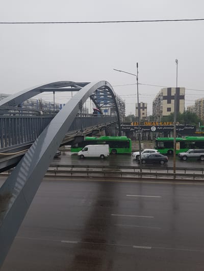 The image depicts a street scene in Бұлақты, Kazakhstan, on a cloudy day. A prominent pedestrian overpass, made of grey metal with arched supports, dominates the foreground. The overpass has a sloped walkway with railings. Below the overpass, a multi-lane road is visible, wet from rain. Several vehicles are on the road, including a white van, a dark sedan, and two silver SUVs. Adjacent to the road and beneath the overpass, a row of green buses, labeled "ECOBUS.KZ," is parked. Behind the buses, a building with signage that reads "IMRAN KAFE" is visible, along with other apartment buildings in the background. A street lamp and utility poles are also present. The overall atmosphere is overcast and damp.