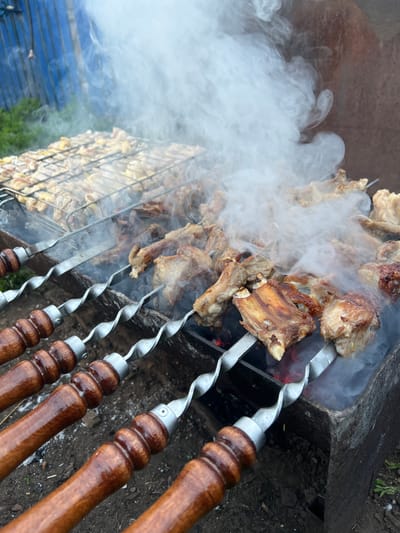 The image captures an outdoor grilling scene in Amirovo, Russia. In the foreground and center, various meats are actively cooking over glowing coals within a rectangular metal grill basin. Several pieces of bone-in meat, likely ribs or chops, are skewered on at least four individual metal skewers, which feature spiral-twisted metal shafts and dark, polished wooden handles. To the left of the skewered meat, a metal grill grate holds smaller cuts of meat, possibly chicken. Thick white smoke rises from the cooking meat, partially obscuring the background. The ground immediately surrounding the grill consists of dark soil. In the background, a blue corrugated metal fence or wall is visible on the left, with sparse green foliage at its base, while a darker, possibly rusted structure appears on the right. No individuals are present in the frame.