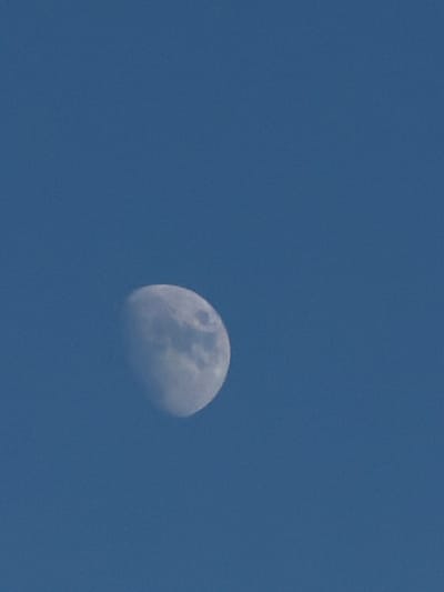 The image shows a portion of the moon against a clear blue sky. The moon is in its gibbous phase, with more than half of its surface illuminated. The illuminated part of the moon displays visible craters and surface features, suggesting it is daytime or twilight. The sky is a uniform shade of blue, with no clouds present. The image appears to be taken from a ground-level perspective, looking upwards. No people, text, or specific location cues are visible in the frame. The overall impression is of a clear sky with the moon prominently displayed.