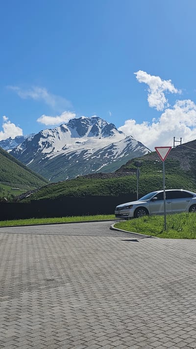A silver sedan is parked on the right side of a paved area, adjacent to a grassy verge, with a mountain range in the background. The mountains are snow-capped and surrounded by green, rolling hills. The sky is blue with scattered white clouds. A triangular yield sign is visible to the right of the car. The paved area is made of interlocking gray pavers arranged in a herringbone pattern. A dark fence runs along the bottom of the green hills. The scene appears to be in a natural, mountainous landscape, possibly Kamskho, Russia, given the context.