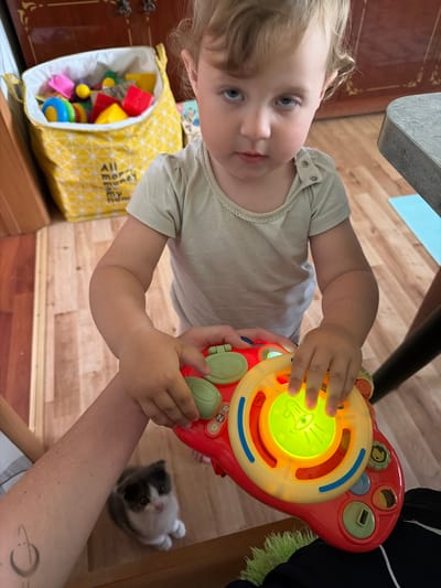 A toddler girl with blonde curly hair and blue eyes is playing with a red and yellow musical toy. She is wearing a light beige t-shirt. Her hands are on the toy, which has colorful buttons and a large illuminated green center. In the background, a yellow fabric bin filled with colorful children's toys is visible. To the left of the girl, a small grey and white kitten is sitting on a wooden floor. An adult's arm with a tattoo is holding the toy from below. The scene appears to be indoors, likely a home environment, with wooden flooring and furniture visible. The location is identified as Komsomolskiy, Russia.