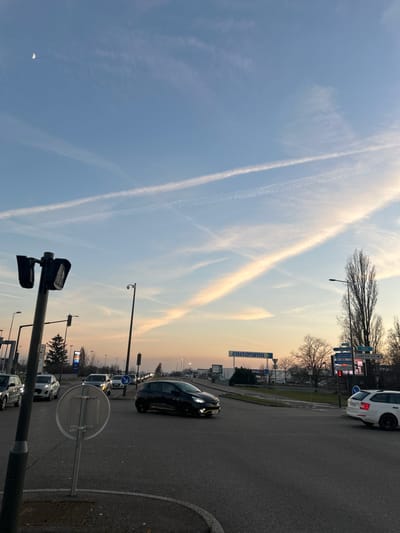 The image depicts an urban intersection in a commercial zone of Vendenheim, France, at twilight. The sky transitions from light blue overhead to orange and pink near the horizon, containing multiple intersecting white contrails. A slim crescent moon is visible in the upper left quadrant.

In the immediate foreground on the left, a black traffic light pole is positioned, supporting two dark-colored traffic light heads. To its right, a white circular regulatory sign with a horizontal dark bar is mounted on a pole.

On the asphalt roadway, a black Renault Clio, identifiable by yellow accents along its lower side panels, is positioned centrally within the intersection with its headlights illuminated, facing generally right. To the left of the intersection, a queue of light-colored vehicles, including white and silver cars, is visible on an approaching lane. On the far right, a white Skoda Octavia Combi is partially visible, positioned at the edge of the intersection.

A tall street light pole, equipped with a surveillance camera, stands to the right of the foreground traffic light. Further right, a multi-directional road signpost displays destinations including "VENDENHEIM" and "LAMPERTHEIM." In the background, a large blue "castorama" sign is prominent on a commercial building, with other indistinct commercial signs visible below it. A line of bare deciduous trees is present along the horizon, behind the commercial structures.