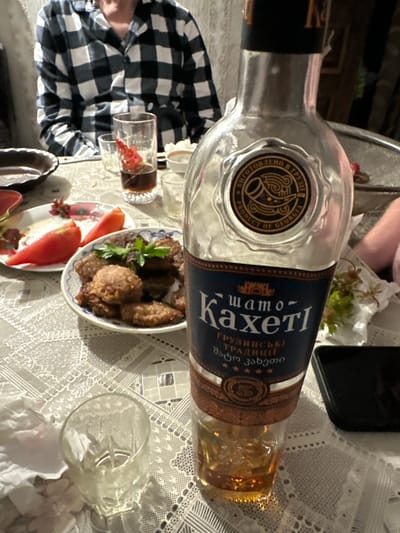 The image shows a table setting with food and drinks, possibly in Vydoshnia, Ukraine. In the foreground, a bottle of "Kakheti" Georgian wine is partially visible, with some amber liquid remaining. Next to it, a small, empty glass with an intricate pattern is placed on a lace tablecloth. A plate with several small, fried food items, garnished with a sprig of parsley, is to the left of the bottle. Further left, another plate contains sliced tomatoes and possibly some berries. A person wearing a black and white checkered shirt is seated in the background. The scene appears to be a casual meal or gathering.