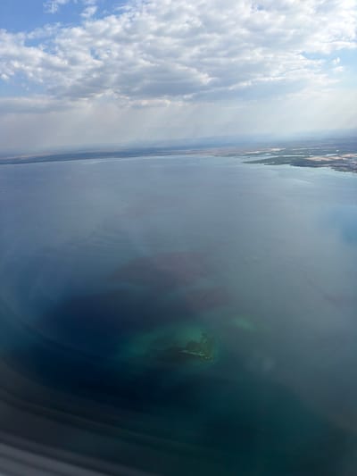 An aerial view captures a coastal landscape under a bright, cloudy sky. The image is oriented vertically, with the clouds occupying the left side and a vast body of water on the right. The clouds are white and fluffy, with some darker, heavier ones suggesting recent or imminent rain, but the overall impression is of a sunny day.

The water is a deep blue-green, with lighter, more translucent areas indicating shallower water or underwater features. A small, rocky island with some vegetation is visible in the water. The shoreline is visible in the distance, with a thin strip of land separating the water from what appears to be drier terrain.

The perspective suggests the photo was taken from an aircraft. The scene conveys a sense of openness and natural beauty. There is no visible text in the image.