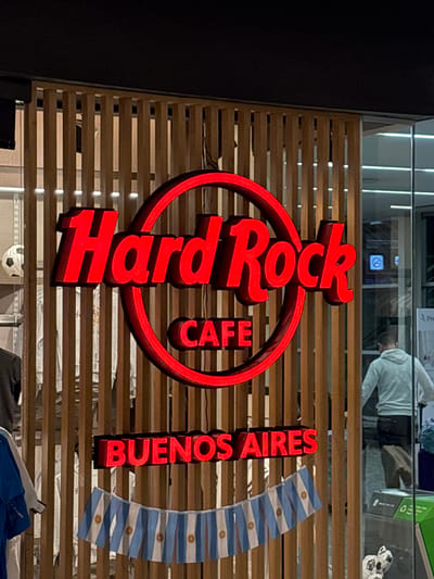 The image shows the exterior of a Hard Rock Cafe in Aeropuerto Internacional Ezeiza, Argentina. A large red neon sign with the words "Hard Rock CAFE" is prominently displayed, centered on a wooden slat wall. Below this, another red neon sign reads "BUENOS AIRES." A string of small Argentinian flags hangs below the "BUENOS AIRES" sign. To the left, partially visible through the wooden slats, is a display with what appears to be merchandise, including a soccer ball and clothing items. To the right, a person wearing a light-colored hoodie and dark pants is walking away from the camera, with luggage visible in the background. The environment appears to be an indoor public space, possibly within the airport terminal.