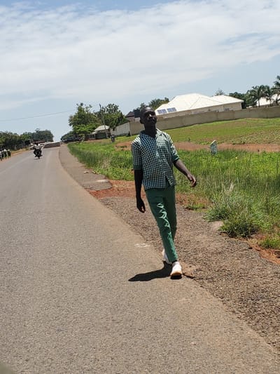 A young Black male, wearing a green and white checkered shirt and green pants with white sneakers, walks along the shoulder of a paved road in Pankshin, Nigeria. He is facing towards the right of the frame and looking upwards and to the right. The road is asphalt, with a gravel shoulder on the right side of the image. Beyond the shoulder is a grassy area with some dry patches and a small white marker post. In the background, a modern house with a white roof and solar panels is visible, surrounded by a low concrete wall. Palm trees and other vegetation are scattered in the background, and a bright blue sky with scattered white clouds dominates the upper portion of the image. Other people and a motorcycle are visible further down the road, suggesting a populated area. The lighting indicates it is daytime, likely mid-morning or afternoon.
