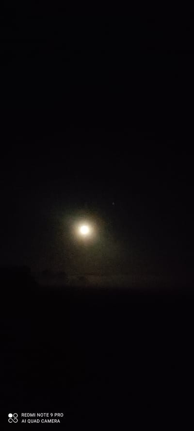 A nighttime photograph captures a bright full moon partially obscured by a thin haze, casting a soft glow over a dark, silhouetted landscape. The moon appears slightly off-center, positioned in the upper left quadrant of the image, with a faint halo of light surrounding it. Below the moon, the horizon is a stark, dark line, with subtle variations in darkness suggesting distant hills or trees. The lower portion of the image is dominated by deep shadows, with no discernible features. In the bottom left corner, the text "REDMI NOTE 9 PRO AI QUAD CAMERA" is visible in white lettering, indicating the device used to capture the image. The overall scene is one of quiet solitude, with the moon serving as the sole significant light source in the otherwise inky blackness. The location is the city of Mliiv, Ukraine.