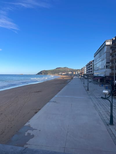 This image displays the beachfront and promenade of Zarautz, Spain, under a clear, bright blue sky with sparse wispy clouds. The scene is bisected by a light gray concrete promenade that runs parallel to the coastline. To the left of the promenade, the Atlantic Ocean features blue-green water with white-capped waves breaking onto a wide, light brown sandy beach. Numerous footprints are visible on the wet sand. In the distance, the beach curves around a prominent green, rolling hill, with indistinct residential or commercial structures visible at its base.

To the right of the promenade, a series of multi-story buildings with light-colored facades and balconies line the street, some appearing to be hotels or apartment blocks. Several ground-floor establishments feature stacked outdoor seating (chairs and tables), suggesting cafes or restaurants. Dark-colored streetlights with spherical, light-colored fixtures are regularly spaced along the promenade's right edge, separating it from the cobblestone-paved area directly in front of the buildings. Three small, indistinct figures are visible walking on the distant sandy beach. A low, dark gray barrier or railing is present in the immediate foreground, at the bottom of the frame. The lighting indicates clear daytime conditions.