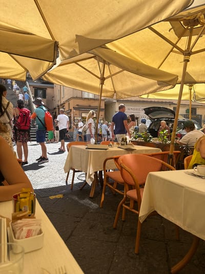 The image captures an outdoor seating area of a cafe or restaurant in Pogerola, Italy, likely on a sunny day. Yellow umbrellas provide shade over tables set with white tablecloths and chairs, some of which are orange and wicker. People are visible in the background, walking up a wide stone staircase and along a street. One person in the foreground is seated at a table, with their arm resting on the table. Another table in the foreground has sugar packets and a glass. The architecture suggests a Mediterranean setting, with stone paving and buildings in the background. A black car with its trunk open is parked on the street, and a sign that reads "FARMACIA" is visible on a building. Another sign above the car reads "ANDREA". The overall scene depicts a bustling daytime atmosphere in a picturesque Italian town.