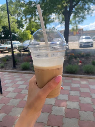 The image shows a close-up of a person's hand holding a clear plastic cup filled with an iced coffee drink. The drink has multiple layers, including coffee, milk or cream, and what appears to be whipped cream or foam on top. A white straw is inserted into the drink. The person's hand has light skin and a French manicure with pink nail polish and white tips.

The background is blurred but suggests an outdoor setting. To the left, there are green trees and a bright blue sky with some clouds, indicating it's daytime. Hints of buildings and vehicles are visible in the distance. To the right, a patterned ground composed of pink and white tiles is visible. The lighting suggests it is a sunny day. The overall impression is a casual moment, likely enjoying a refreshing beverage outdoors on a pleasant day. There is no visible text in the image. The context provided places the scene in Ivanivka, Ukraine.