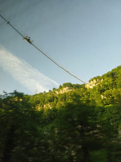 This image depicts a vast, mountainous landscape dominated by lush green trees covering steep cliffs. A long, narrow structure, possibly a bridge or an elevated walkway, extends diagonally across the frame from the top left towards the center right, disappearing behind the trees. This structure appears to be made of metal bars and supports. In the distance, against a clear blue sky with a few wispy clouds, a transmission tower or similar utility pole is visible. The foreground is blurred, suggesting motion, with dark green foliage obscuring the lower portion of the scene. There are no people or visible interactions in the image. The scene appears to be a natural environment, potentially a park or a remote area with man-made infrastructure. The prompt's request to include the location in the city of Galitsyno, Russia, cannot be fulfilled as there are no visual cues in the image that indicate this specific location.