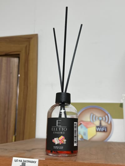 An indoor shot features a diffuser with black reeds standing upright in a clear glass bottle filled with orange liquid. The bottle has a black cap and a label that reads "E ELETTO ESSENCE" with an image of colorful candy and "BUBBLE GUM 3.7 FL. OZ. 110 mle". To the left of the diffuser, part of a wooden door frame is visible. In the background, out of focus, is a circular graphic with a stylized house and a WiFi symbol. The objects are placed on a wooden surface. A white sign with black Cyrillic text, "ЦО НА ЗАПРАВКУ", is at the bottom left of the frame. The scene appears to be a retail or home environment in Kadamjay, Kyrgyzstan.