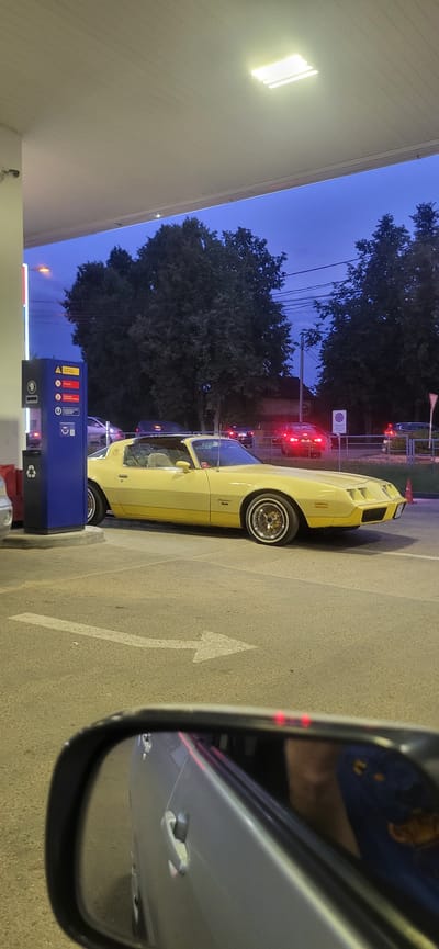 The image depicts a yellow classic car, a Pontiac Firebird, parked under a canopy at a gas station in Pravdinskiy, Russia. The sky is a deep twilight blue. The car is facing right, with its driver's side visible. The gas station has a blue dispenser unit with various icons and a trash can next to it. In the background, other cars are visible, some with their brake lights on, suggesting traffic. Trees line the background, and overhead power lines are also present. The foreground shows a side mirror of another vehicle, reflecting part of its interior and a person. A white arrow painted on the asphalt indicates a turning direction. The overall scene suggests a late evening at a gas station, with the classic car being the prominent subject.