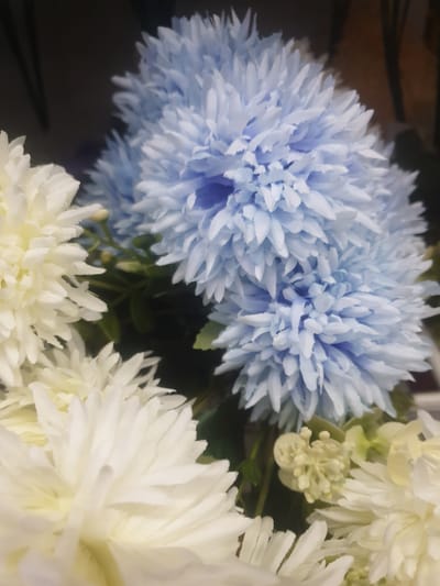 The image displays a close-up view of artificial flowers, predominantly in shades of light blue and white. The flowers have a layered, spiky petal structure. The background is dark and out of focus, with hints of brown and dark green. The scene is an indoor setting, possibly a display or decoration. There is no indication of a specific location within Kauman, Indonesia. The image does not contain any people or actions.
