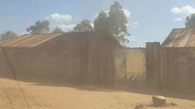 A dusty landscape reveals two weathered wooden structures with corrugated metal roofs, partially obscured by haze. One building displays a door and a small window. A corrugated metal gate stands between the buildings. Trees and a pale sky with light clouds are visible in the background. The ground is covered in dirt.