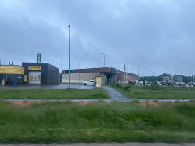 The image depicts a commercial area in Harkujärve, Estonia, on an overcast day. In the foreground, blurred green grass with some red markings is visible. In the midground, a row of buildings includes a dark-colored structure with a yellow sign displaying "AUTOPESU" (car wash in Estonian) and a white garage door. Adjacent to it is a larger building with a brown, slatted facade, bearing a green "PRISMA" logo. A white car is parked near a yellow vending machine outside this building. Further down the street, several cars are parked and driving, and there are apartment buildings in the background. Streetlights are visible throughout the scene.