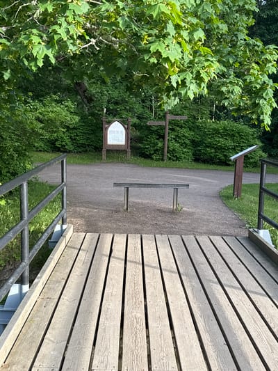 The image depicts an outdoor scene at Keila-Joa Park in Keila-Joa alevik, Estonia. In the foreground, a wooden footbridge with light gray planks leads towards a gravel path. Black metal railings line the sides of the bridge. The path curves gently to the right and is bordered by green grass and dense foliage.

Further into the scene, a simple wooden bench with a dark gray metal seat is positioned on the path. To the left, a brown wooden signpost with an arched top displays text and an illustration. Adjacent to it, another wooden signpost indicates directions with two horizontal arms. To the right, a narrower brown wooden post stands at an angle, topped with a slanted information board. The background is filled with lush green trees and bushes, suggesting a park or natural setting. The lighting indicates it is daytime, with sunlight casting shadows. No people are visible in the image.
