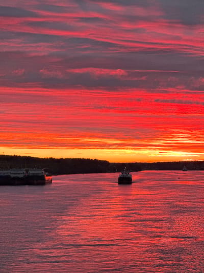 A dramatic sunset over the Svir River near Voznesenye, Russia, paints the sky with vibrant bands of red and orange. Wispy, horizontal clouds are illuminated by the setting sun, creating a fiery spectacle. The river's surface reflects the intense colors of the sky, appearing as a shimmering expanse of pink and red.
In the foreground, a large barge or freighter is docked on the left side of the image, its dark hull contrasting with the bright water. Further out on the river, a smaller vessel, possibly a tugboat, is visible in the center, heading towards the viewer. In the distance, along the far bank of the river, a dense line of dark trees forms a silhouette against the colorful horizon. A few smaller boats or buoys are scattered in the water. The overall scene is one of natural beauty and tranquility, with the river and sky dominated by the rich hues of sunset.