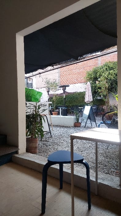 The image depicts an outdoor patio or courtyard viewed from an interior threshold. The foreground shows a light-colored floor, a dark blue four-legged stool, and the edge of a light-colored table. A concrete ledge separates this interior space from the outdoor area. The outdoor patio, located in Tlaxcala City, Mexico, is surfaced with light-colored gravel and partially covered by a dark mesh shade cloth supported by a metal frame.
Within the patio, a large potted plant in a terracotta pot is positioned on the left, alongside a white counter-like structure topped with green material and a black A-frame sign. A tall, dark patio heater stands further back. In the center, a low, light-colored concrete barrier or planter contains smaller potted plants. To the right, a folded light-colored market umbrella is visible, accompanied by another black A-frame sign and a black and blue woven chair. The background features a reddish-brown brick wall, with parts of a light-colored urban building and sparse green foliage visible above and behind it. No individuals are present in the scene.