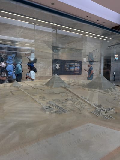 In Kafrat Nassar, Egypt, a detailed architectural model of the Giza pyramid complex is displayed under glass. Three pyramids, representative of those at Giza, are prominent features of the model, which also includes smaller structures and pathways. The sand-colored model is illuminated by overhead lighting, visible as reflections on the glass display case. In the background, several people are reflected in the glass. One person, a woman wearing a blue cap and white shirt, holds a water bottle. Another person, a man in a grey t-shirt and shorts, looks at a tablet. A group of individuals, including a woman in a floral shirt, a man in a blue shirt with a patterned scarf, and another man in a teal shirt, are also visible in the reflection, appearing to be observing the model. The environment behind the glass features light-colored walls and suspended lighting fixtures.