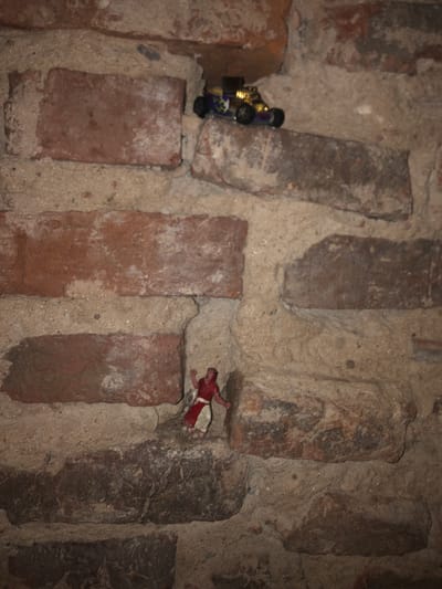 The image depicts a close-up view of a brick wall. A toy car sits on top of a brick on the left side of the image, while a doll is positioned between two bricks on the right. The mortar between the bricks is a light color. The lighting is dim. The location of Beccar, Argentina, is speculative.