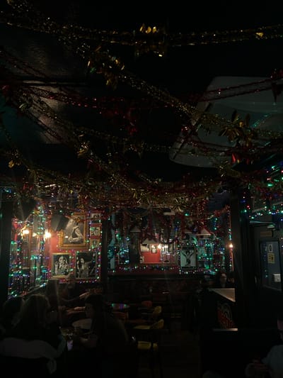 A dimly lit interior is decorated with garlands and twinkling lights. Reflected in mirrors are framed photos, creating a seemingly infinite gallery. A large, white rectangular object hangs near the ceiling on the left. People are visible on the right side of the image, but they are not in focus. The scene feels festive and slightly crowded.