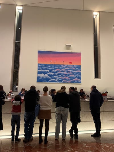 A group of people stand in a room, some looking up at a large art piece hanging on the wall. The artwork is a stylized image of a sunset over clouds. The room has high ceilings, with lights recessed into the ceiling. Long windows are visible on the left side of the frame. The floor is a reflective surface, mirroring the people standing on it.