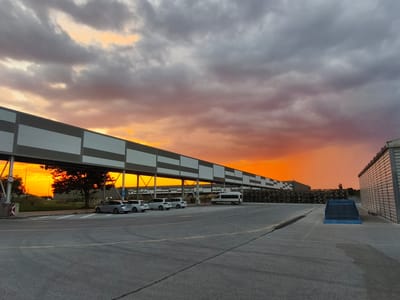 This media file captures an industrial scene at dusk in Benkovski, Bulgaria. The setting appears to be a large facility, possibly a logistics or manufacturing site, characterized by a long, elevated walkway or conveyor belt system running horizontally across the frame. This structure is composed of grey and white rectangular panels.
Below the walkway, several white SUVs and a white van are parked in a paved lot. In the distance, stacks of what appear to be building materials or equipment are visible, stretching along the base of the walkway. To the right, a building with a corrugated facade stands prominently.
The most striking element of the image is the dramatic sunset. The sky is filled with dark, heavy clouds that are illuminated from below by a vibrant, fiery orange and yellow glow, suggesting the sun is low on the horizon. This creates a stark contrast between the dark, utilitarian structures and the intense natural light. The overall mood is one of quiet transition from day to night, with a sense of vastness conveyed by the expansive sky.
A sign with "5,5" is visible on a pole near the left edge of the frame, likely indicating a speed limit or height restriction. There is no visible text on the buildings or vehicles. The lighting and cloud formations indicate it is late evening, shortly after sunset. The weather appears to be clear with some cloud cover.