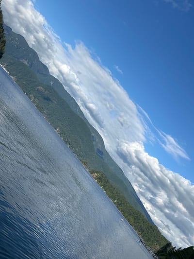 The image depicts a panoramic view of a body of water with mountains in the background. The scene is captured from a low angle, likely from a boat on the water.
In the foreground, the water is calm with gentle ripples, reflecting the blue sky. The color of the water is a deep blue, suggesting depth.
The middle ground is dominated by a lush, green mountainside that slopes down towards the water. The vegetation appears dense, consisting of trees and shrubs. Scattered along the base of the mountain, near the water's edge, are several small white structures, likely houses or cabins, indicating a residential area.
The background features a series of imposing mountains, layered and receding into the distance. Their peaks are partially obscured by a dynamic sky filled with a mixture of bright white cumulus clouds and wispy cirrus clouds, interspersed with patches of clear blue sky. The angle of the photograph, with the horizon tilted diagonally, adds a sense of dramatic perspective.
The presence of a pier or dock structure can be observed along the shoreline in the lower right portion of the image, with some small objects that could be boats or watercraft nearby. A tall transmission tower is also visible on the mountainside in the distance.
This scene is consistent with the geography of the District of North Vancouver, Canada, particularly areas along the shores of Indian Arm or Burrard Inlet, known for their mountainous terrain and waterfront communities.