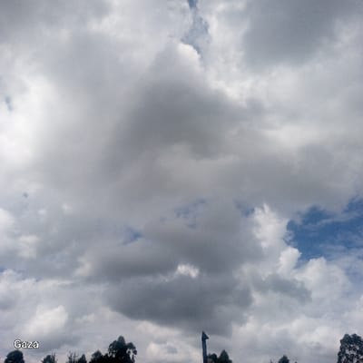 This image depicts a predominantly cloudy sky over Bonyamaro ward, Kenya. The sky is filled with a dense formation of stratocumulus clouds, exhibiting various shades of grey and white. Multiple patches of clear blue sky are visible through breaks in the cloud cover, primarily towards the right side of the frame. At the bottom of the image, a treeline with dark green foliage is visible, with individual tree crowns distinguishable. Emerging from behind the treeline, slightly right of the bottom center, is a dark, pointed, man-made structure, possibly a tower or spire, which is partially obscured. No people, animals, specific actions, or interactions are discernible. The word "Gaza" is visible in white text in the lower-left corner.