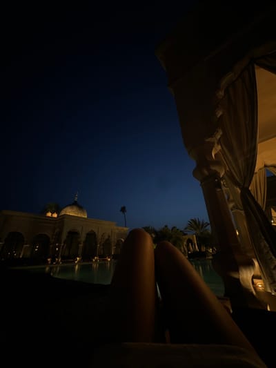 The image shows a pair of tanned legs, likely a woman's, resting on a lounge chair. The person is lying on their back, facing the camera, with the top half of their body out of frame. The scene is at night, with a clear, dark blue sky and the silhouette of a building illuminated by artificial light. There are palm trees and other vegetation in the background, suggesting a resort or hotel setting. The location, at coordinates 31.663, -7.872, corresponds to a secluded luxury resort in the outskirts of Ouled Jelal, Morocco. The scene is calm and peaceful, with a relaxed ambiance. The person is likely enjoying a moment of tranquility.