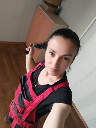 A woman stands in a room, holding one of her braided pigtails. She is wearing red overalls over a black t-shirt and a gold necklace with a cross pendant. Her hair is dark and pulled back. The room has wooden flooring and light-colored walls. A peach-colored cabinet with two doors is visible in the background, as well as some cardboard boxes on top of it. The environment suggests an indoor setting, potentially a home or workshop. There are no identifiable landmarks linking this scene to Târgu Jiu, Romania.