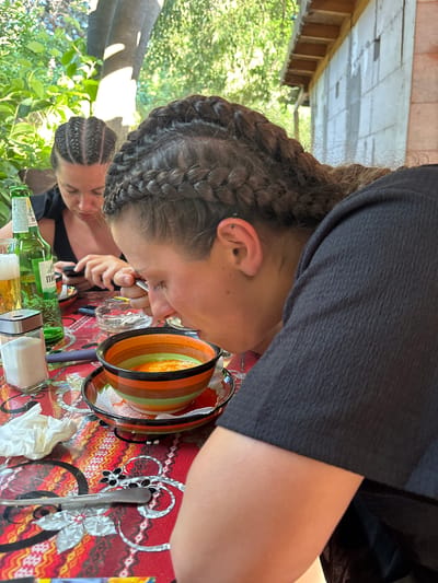 This image depicts two individuals seated at a table outdoors, likely in Kresna, Bulgaria, given the context. In the foreground, a person with braids is leaning over a colorful bowl, eating soup with a spoon. Their face is partially obscured by their head as they focus on their meal. To the left, another person with braided hair is visible, looking down at their phone. On the table, there's a green bottle of beer, a glass of beer, a salt shaker, a small ashtray, and a crumpled napkin. The table is covered with a red and white patterned tablecloth. In the background, lush green foliage and a partially constructed building with concrete blocks are visible, suggesting a casual outdoor dining setting.