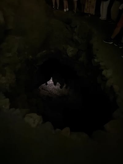 The image shows the entrance to a cave in Haría, Spain. The entrance is dark and surrounded by rocks. A group of people can be seen standing at the top of the entrance. Their feet and shoes are visible at the upper left of the image. Light penetrates the cave interior from the opposite end.
