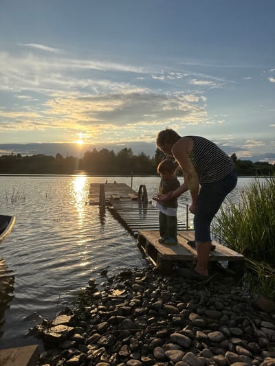 A woman and a young child stand on a wooden dock at the edge of a body of water, likely a lake or river, in Sitno, Russia. The sun is setting, casting a warm glow across the scene and reflecting on the water's surface. The sky is a mix of blue and white clouds, with the sun partially obscured by them. The dock is constructed from weathered wood and extends out over the water. The child, facing away from the camera, appears to be looking at something in the woman's hand. The woman is bent over, interacting with the child. The shoreline is lined with rocks and tall grasses. The overall atmosphere is peaceful and serene, with a hint of familial connection.