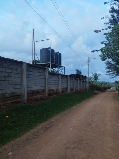 The image shows a dirt road leading towards a concrete wall topped with a metal fence.  Two black water tanks are on top of a metal structure behind the wall. The wall is constructed with interlocking concrete blocks and is approximately five to seven feet in height. The road is dusty and appears to be sparsely used. There are no people or vehicles visible in the scene. It is possible that the road is a small, rural path. The image was captured in the late afternoon, as indicated by the shadows in the scene. The location of the image is Othaya, Kenya, as indicated by the coordinates, which correspond to a residential area on the outskirts of the town. The vegetation is typical of the region, with short, green grass and a few trees visible in the background.  The sky is blue and cloudy, and there are power lines running overhead. There is a streetlight on the right side of the road.  The image does not depict any specific event or action. It is simply a documentation of the scene as it appears.