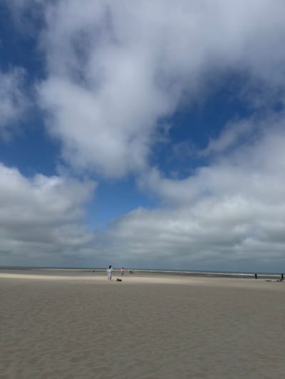 The image depicts a wide, sandy beach at Berck, France, under a sky filled with a mix of blue patches and dramatic, puffy clouds. The sand in the foreground is textured with ripples, suggesting the tide has recently receded. In the middle ground, several figures are visible. A person in a white outfit walks across the sand, while another person in pink is further to the right. Several other individuals and small groups are scattered along the shoreline and in the distance, with one person appearing to be standing near a blue object that could be a surfboard or kite. The sea is a muted gray-blue, meeting the horizon under the cloudy sky. The overall impression is of a tranquil, somewhat breezy day at the beach.