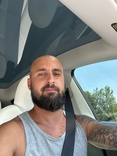 A bald, middle-aged man with a full beard, wearing a gray tank top and a black seatbelt, is seated in the driver's seat of a car. The car has a white interior and a panoramic glass roof. The man is looking at the camera. Outside the car, trees and a bright blue sky are visible through the window. The image was taken in Aurora, United States of America.