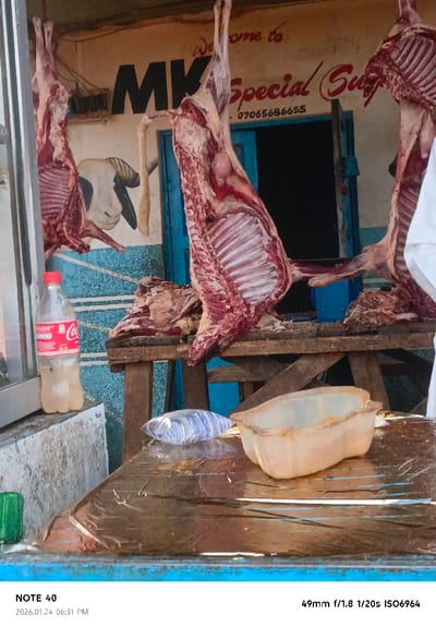 A butcher shop is captured with hanging animal carcasses on display. A painted sign above reads "Welcome to MK Special Su" followed by a phone number. Below the meat, there's a wooden table cluttered with plastic containers and packaging. A Coca-Cola bottle sits near a window to the side. The image appears to be taken in front of the shop, focusing on the display of meat and the business signage.