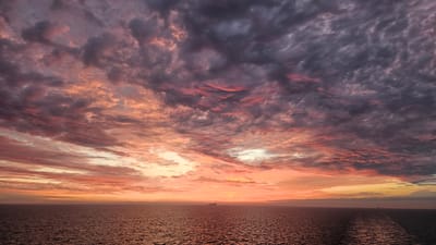 The image depicts a dramatic sunset over a vast expanse of ocean. The sky is filled with a complex tapestry of clouds, painted in vivid hues of orange, pink, and purple. Some clouds are dark and ominous, while others are illuminated by the setting sun, creating a striking contrast. The ocean below reflects these colors, with the water appearing a deep, rich red-brown in the foreground and gradually lightening towards the horizon. A solitary cargo ship can be seen in the distance, a small silhouette against the vibrant sky, further emphasizing the scale and emptiness of the seascape. The horizon line is sharp and clear, dividing the tumultuous sky from the calm water. There are no visible signs of land or urban structures, suggesting a remote oceanic setting, possibly in the open sea far from any coastline. No people or specific landmarks are identifiable in the scene.