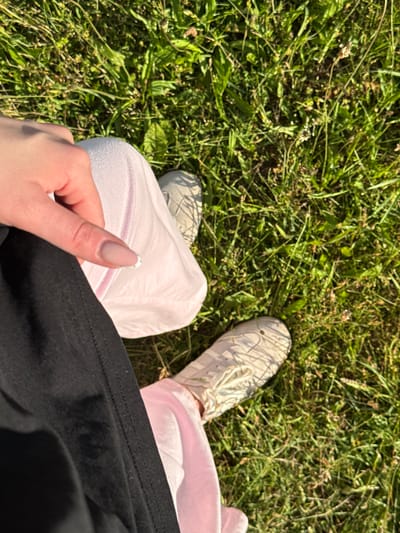 The image shows a person's legs and hand on a grassy area. The person is wearing a black skirt and a light pink garment, possibly a dress or skirt, with white sneakers. The hand is resting on the pink garment, and the fingernails are manicured with a light pink and white design. The background consists of green grass and some small plants. The lighting suggests it is daytime, possibly late afternoon due to the long shadows cast by the grass. The scene is outdoors, on natural ground. There are no discernible landmarks or features that would pinpoint the location to Ivatsevichy, Belarus.
