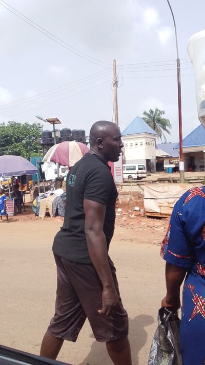 The image depicts an active street scene in Igbo Ukwu, Nigeria, under an overcast sky. In the foreground, a bald, dark-skinned male wearing a black short-sleeved t-shirt with light-colored text on the back and dark brown shorts walks from right to left, his mouth slightly open. His left arm is bent, and his hand is partially clenched. To his right, partially visible, is a dark-skinned female wearing a blue dress patterned with red and white designs, holding a metallic silver and black patterned bag in her left hand.

The street surface is light brown and appears paved. In the mid-ground on the left, an open-air market is visible with multiple vendors under colorful umbrellas (one maroon and beige striped, one purple), displaying clothing items. Behind these stalls, a structure supports several black water tanks and a solar panel street light. Further back on the left, green foliage is present, and a yellow tricycle (keke Napep) is partially visible.

In the mid-ground on the right, several white buildings with distinctive blue pyramidal roofs are present. One building appears to be a utility structure, possibly a power transformer station, with horizontal vents. Another building displays a partially legible sign including text like "SIGN BOARD," "LAUNDRY," and "DRY CLEAN." A white van or mini-bus is parked between these buildings. A palm tree is visible behind the buildings on the right.

Tall utility poles, one wooden with multiple wires and another metal with a street lamp arm and a white translucent container, extend into the upper frame. The ground in the mid-ground features exposed reddish-brown earth and scattered debris.