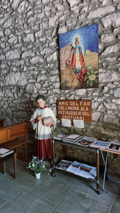 The image shows an interior scene within a stone-walled room, likely a sanctuary or historical site in Susqueda, Spain. In the center-left, a life-sized statue of a young boy dressed in religious attire, including a white surplice with red trim and blue ribbon, stands holding a small wooden box. To the right of the statue, a wooden sign with text in Catalan reads "AMIC DEL FAR COLLABORA A LA RESTAURACIÓ DEL SANTUARI" (Friend of the Lighthouse, Collaborate on the Restoration of the Sanctuary). Below this sign, two smaller sheets of paper are attached, each with the text "LLIBRES DONATIU LA VOLUNTAT" (Books, Donation, Voluntary).

Above the wooden sign, a framed painting or print is hung on the stone wall. It depicts a religious figure, possibly the Infant Jesus of Prague, wearing a crown and ornate robes, standing in a landscape with mountains and flowers. The painting has text at the bottom, including "GRÀCIES NEN JESÚS DE PRAGA" (Thanks Infant Jesus of Prague).

To the left of the statue, a wooden pew or bench is visible. In front of the statue and to the right, a metal cart with wheels holds stacks of books and magazines. One of the magazines visible is titled "Catalunya Cristiana". A white vase with a bouquet of artificial flowers sits on the floor between the statue and the cart. The floor is tiled with a gray, patterned surface. The overall impression is a small, dedicated space possibly within a pilgrimage site or church, with elements of art, devotion, and information for visitors or donors.